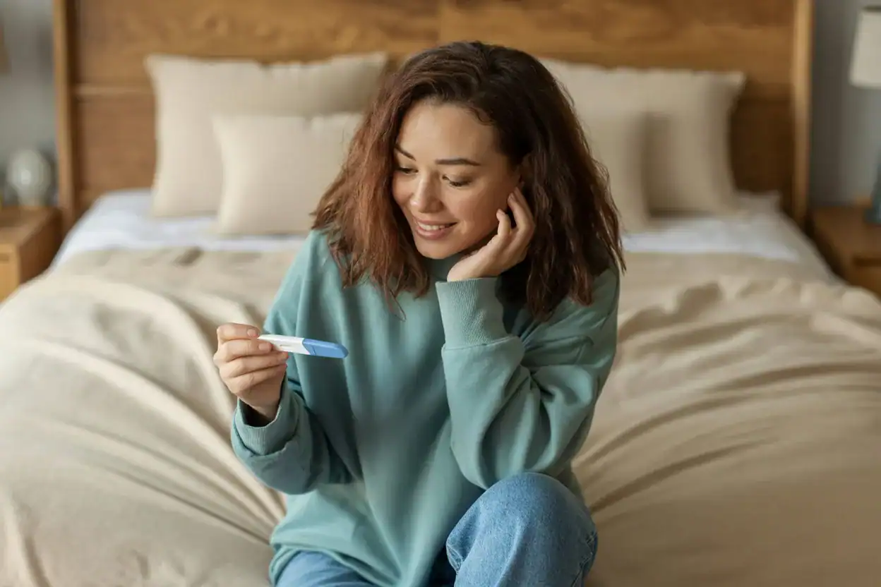 Young woman sitting on a bed in natural light, smiling while holding a positive pregnancy test, wearing a teal sweatshirt and jeans, with soft beige bedding in the background