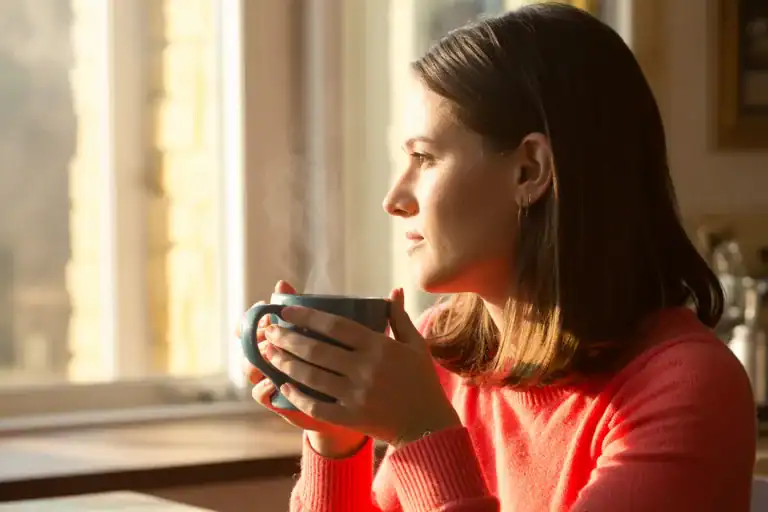 Woman in a coral sweater holding a steaming mug and looking pensively out a window, sunlight illuminating her face in a calm, reflective moment